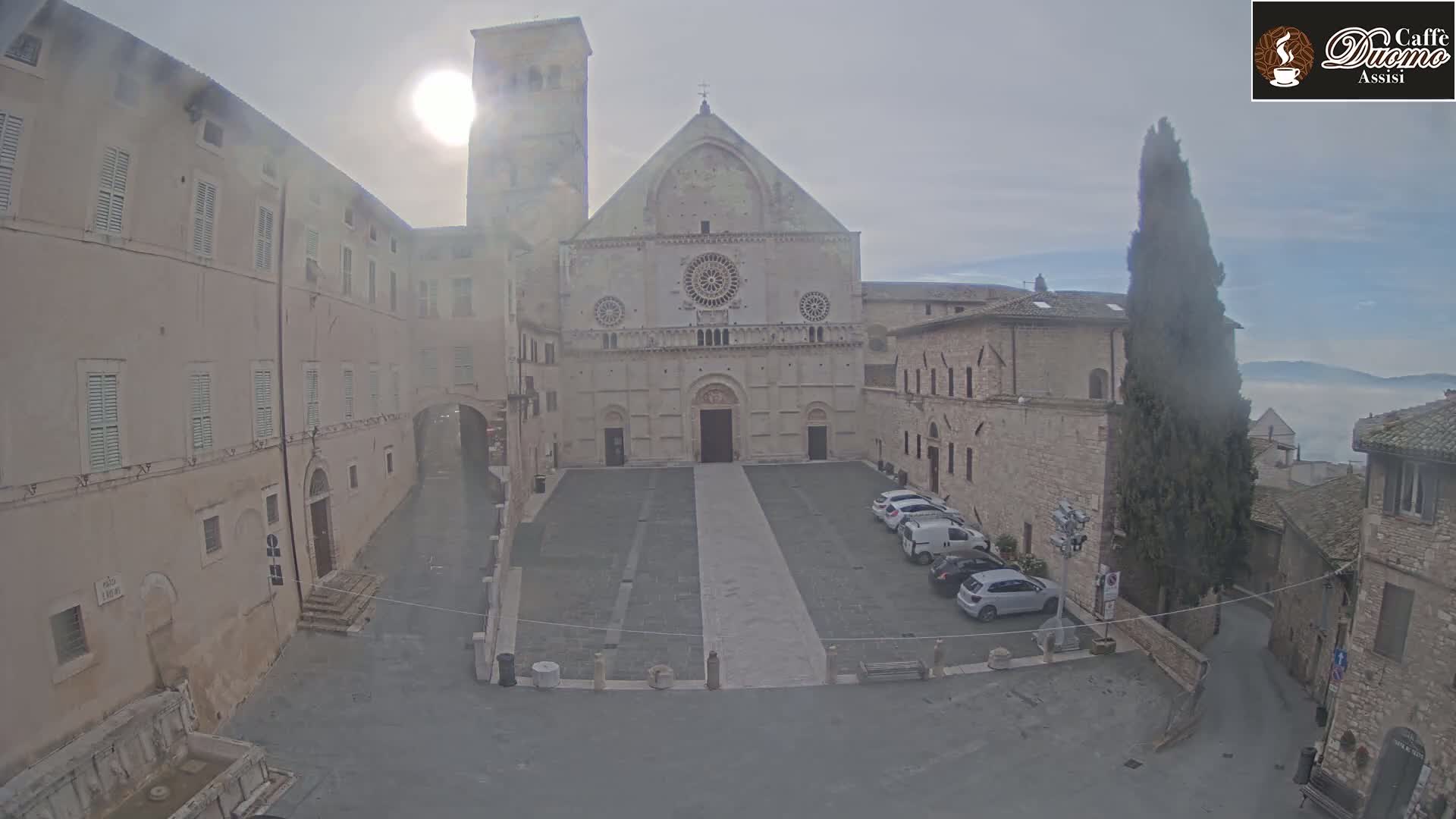 A large stone cathedral sits in a plaza surrounded by buildings on an overcast day with a hazy background.