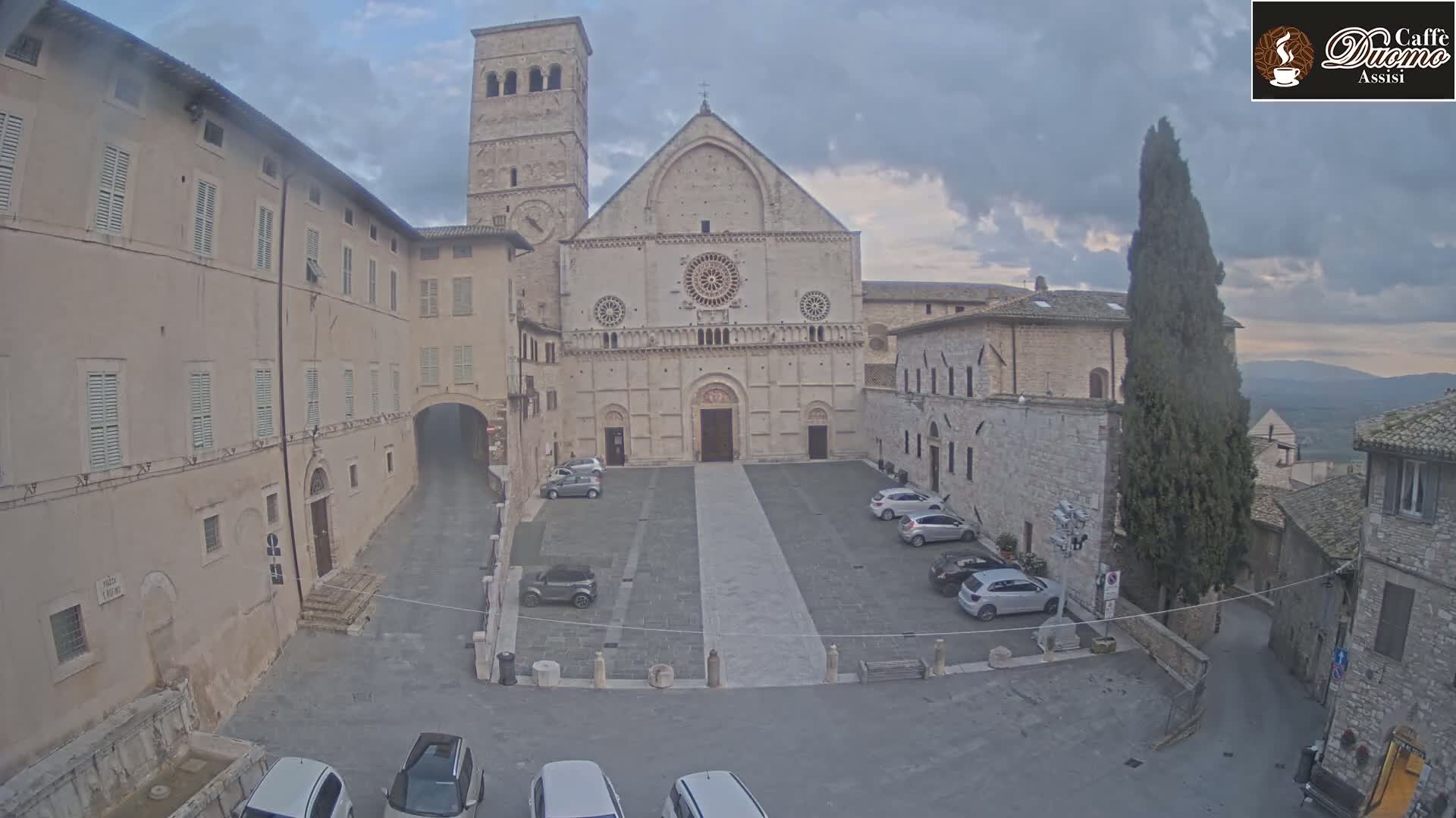 A stone cathedral and surrounding buildings, some with parked cars in front, sit in a town square under a cloudy sky.