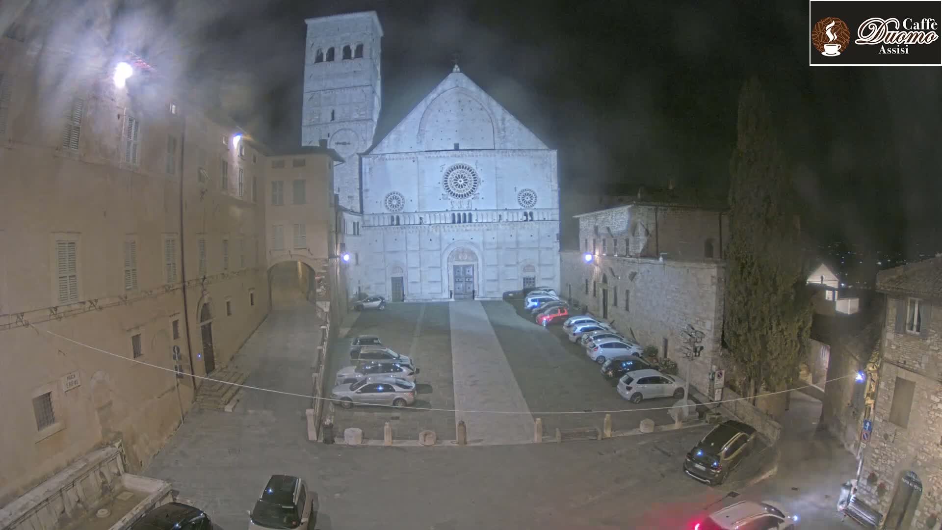 At night, under a hazy sky, a large stone cathedral stands in a town square with cars parked around it.