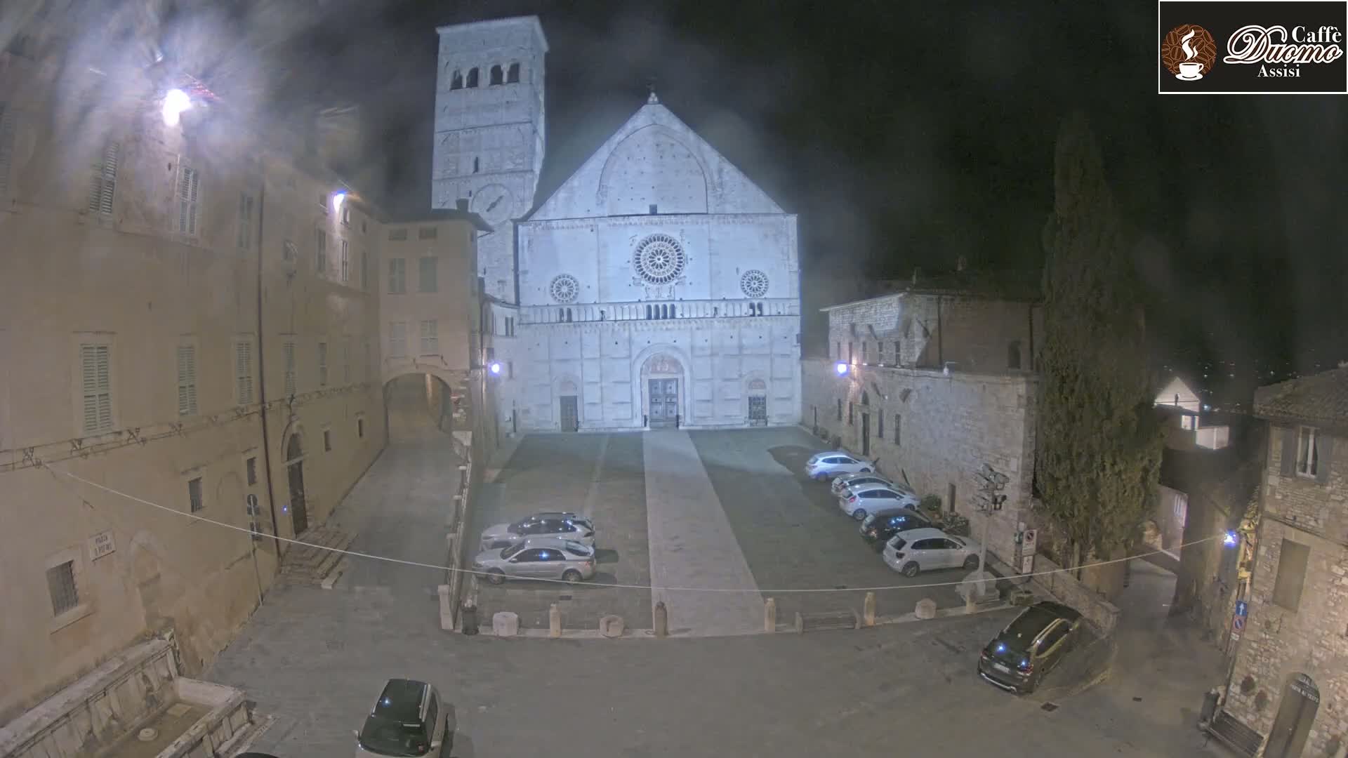 A nighttime view of a large stone cathedral and surrounding buildings in a town square with several cars parked, under a slightly hazy sky.