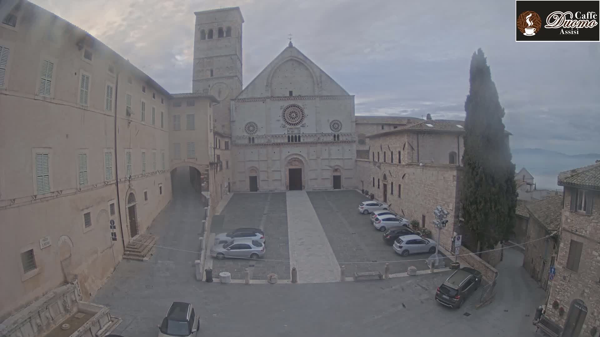 A large stone cathedral stands in a square with several cars parked, under an overcast sky.