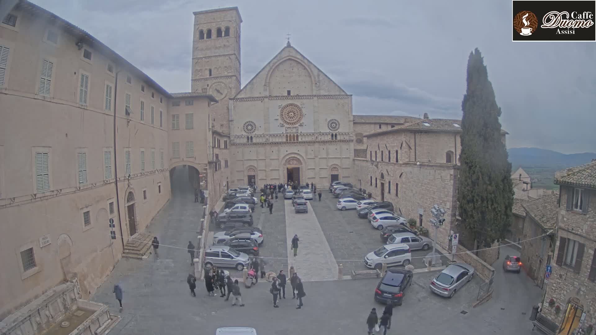 A large stone cathedral stands in a town square, surrounded by buildings, parked cars, and several people; the weather is overcast.