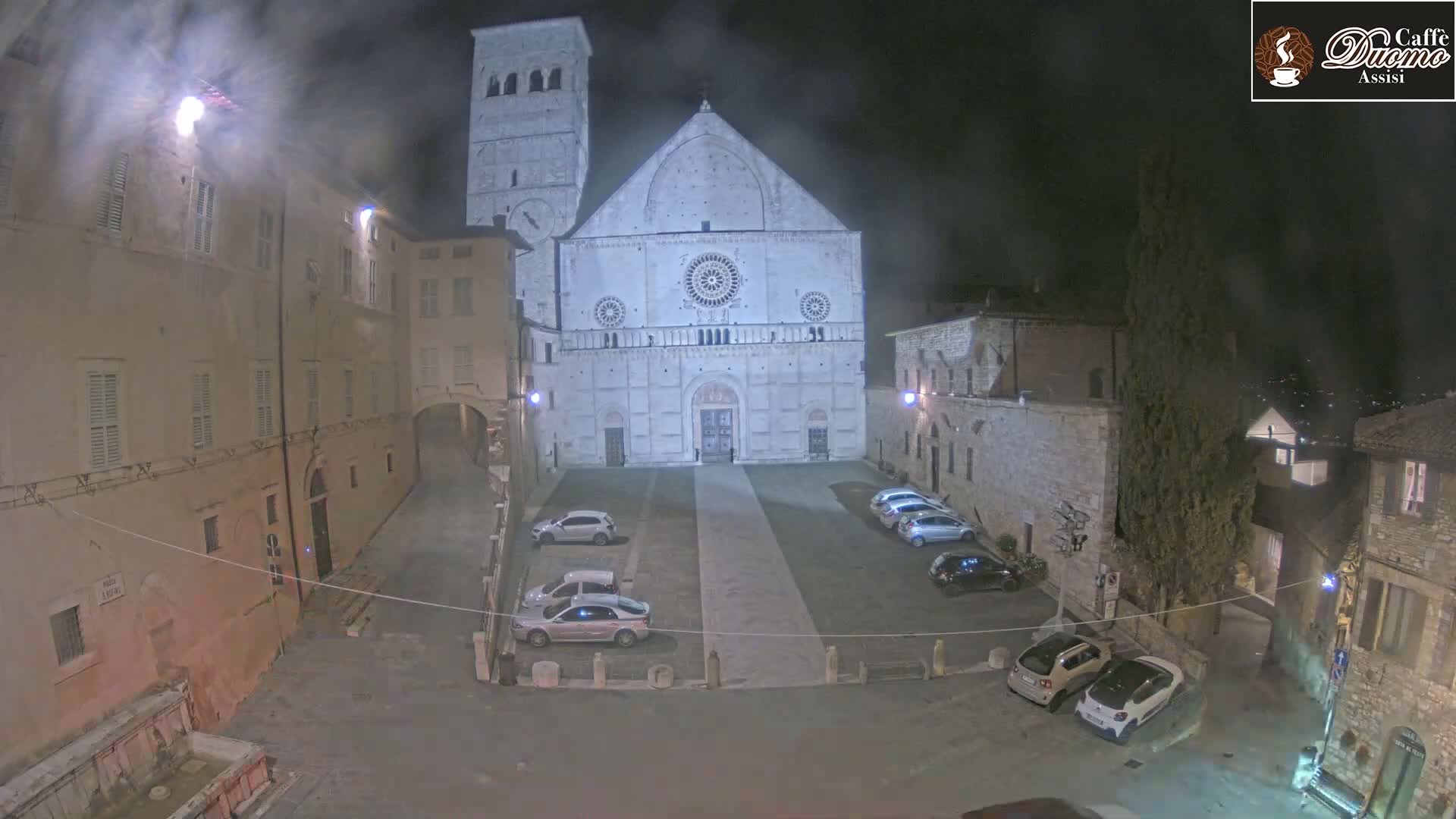 A large, light-colored cathedral stands in a dimly lit town square at night, surrounded by parked cars and older buildings under hazy conditions.