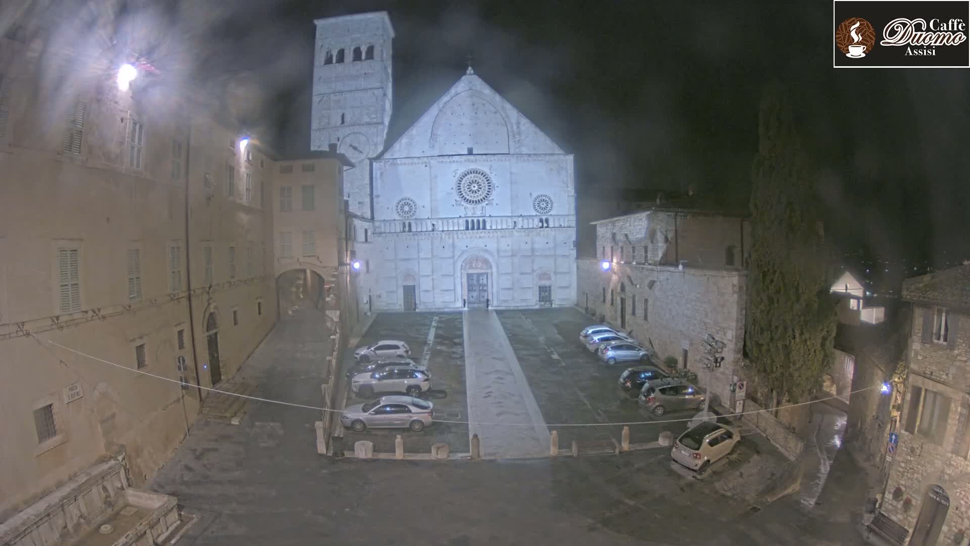 A nighttime view of a large stone cathedral in a town square with several cars parked, under hazy or foggy conditions.