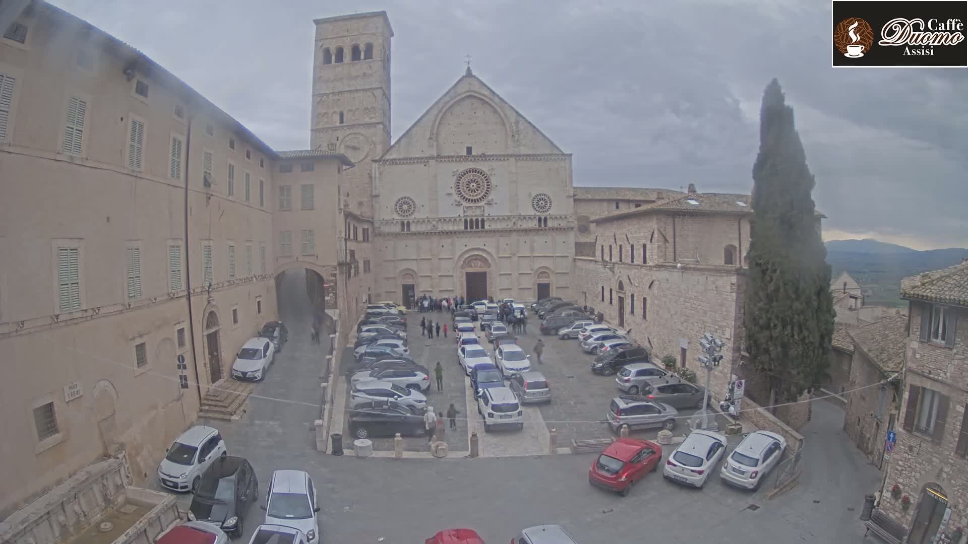 A large stone church with a courtyard filled with parked cars is seen on an overcast day.