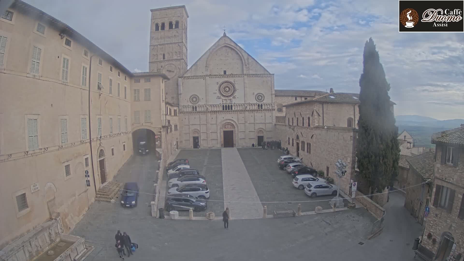 A large stone cathedral sits in a town square with cars parked and several people walking around on an overcast day.