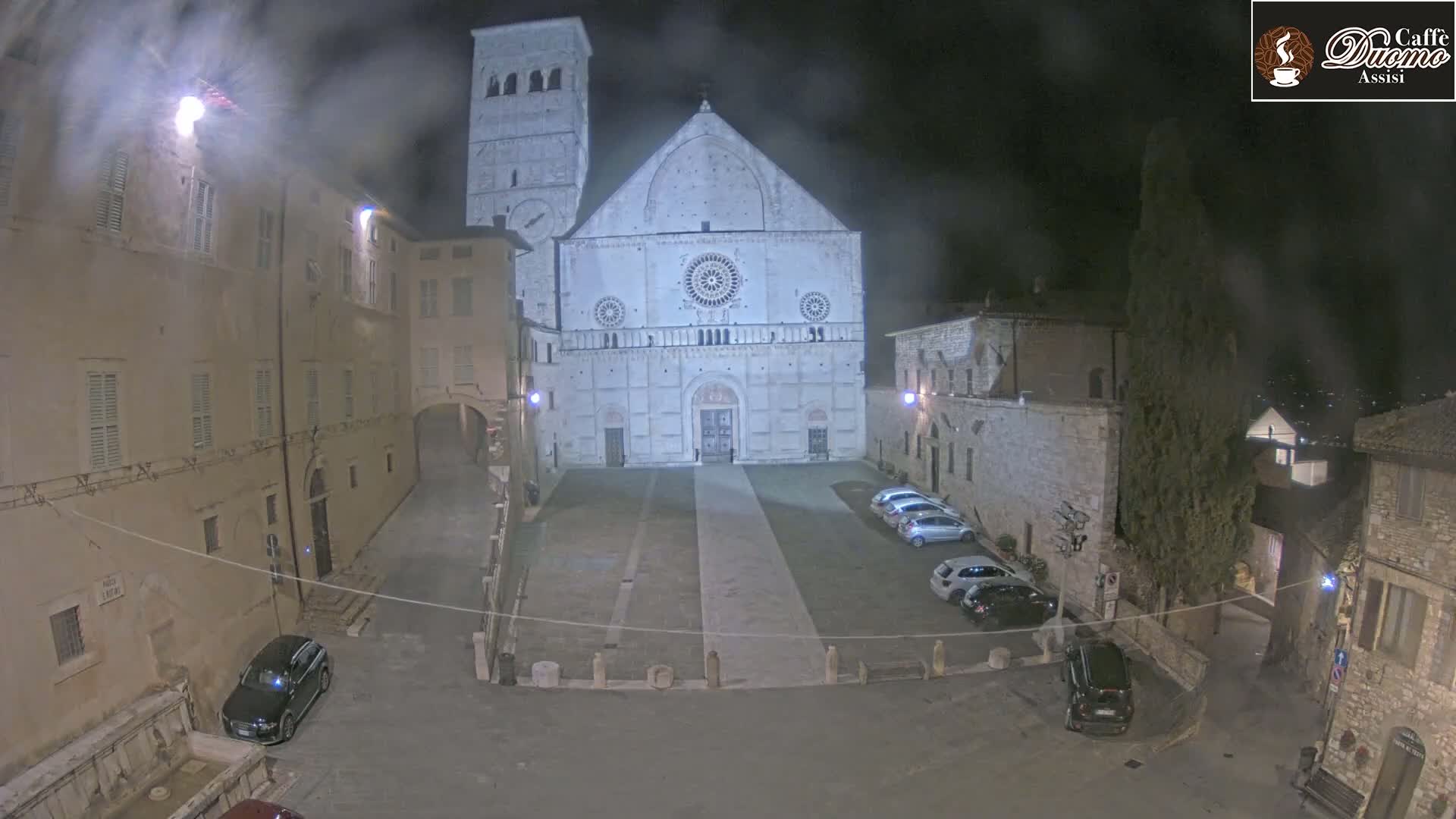 At night, under hazy conditions, a large stone cathedral stands in a town square with several cars parked nearby, surrounded by old stone buildings.