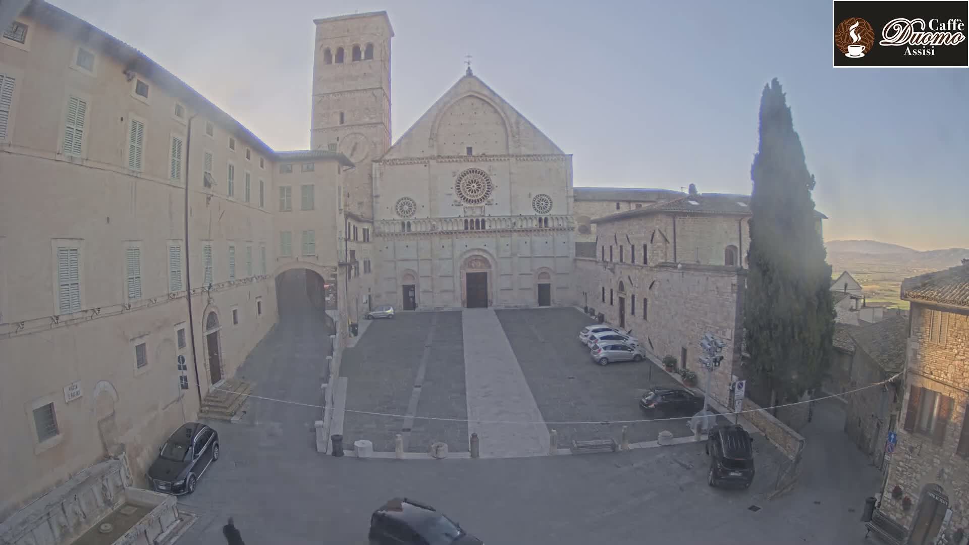 A large stone cathedral stands in a plaza surrounded by older stone buildings, under a mostly clear sky.