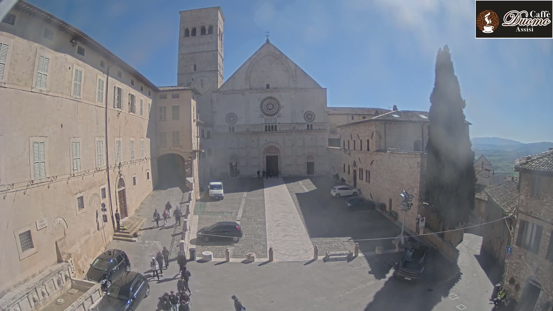 A large stone cathedral and surrounding buildings are visible on a sunny day in a town square with several cars and people walking around.
