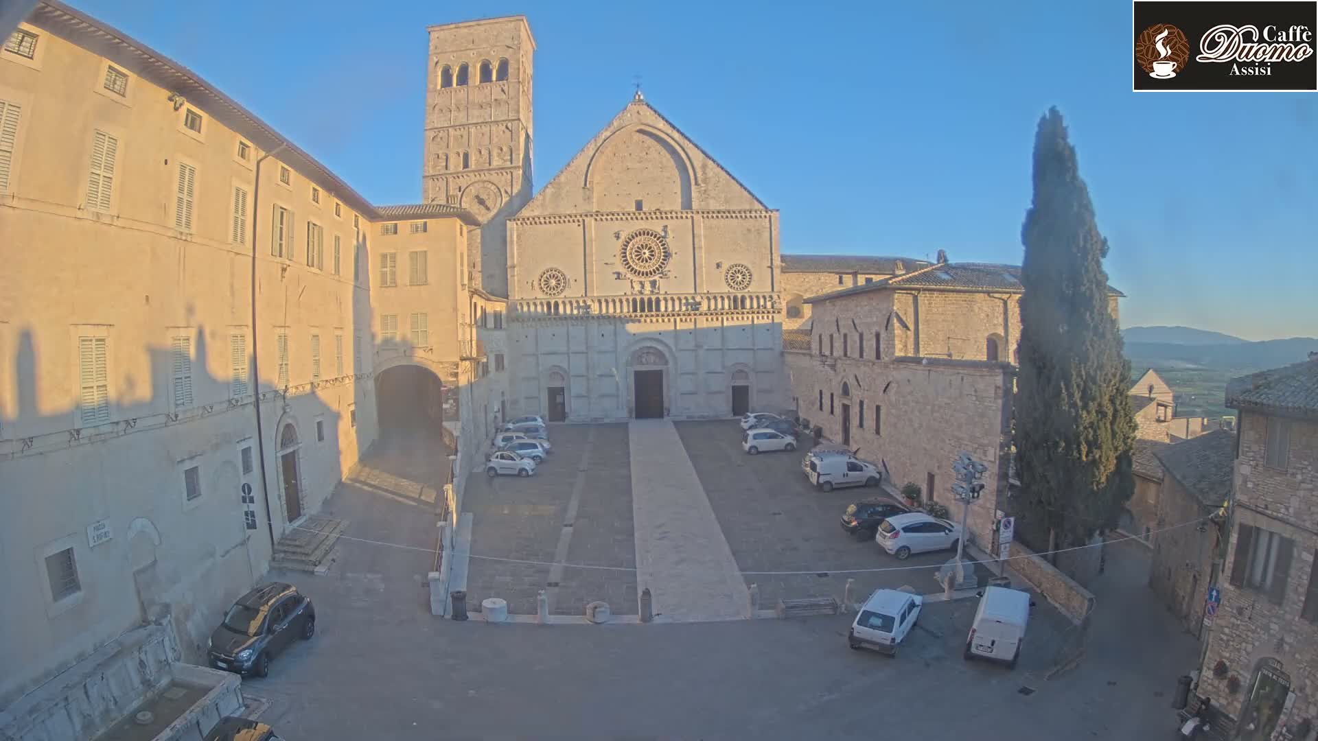 A sunlit stone plaza features a large cathedral with a tall bell tower, surrounded by older buildings under a clear sky.