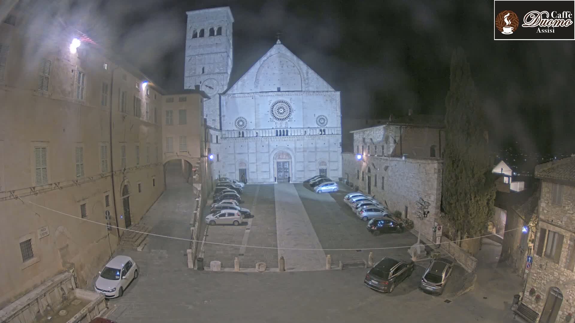 A nighttime view of a town square with a large cathedral at its center, several parked cars, and buildings lining the square under hazy or foggy conditions.