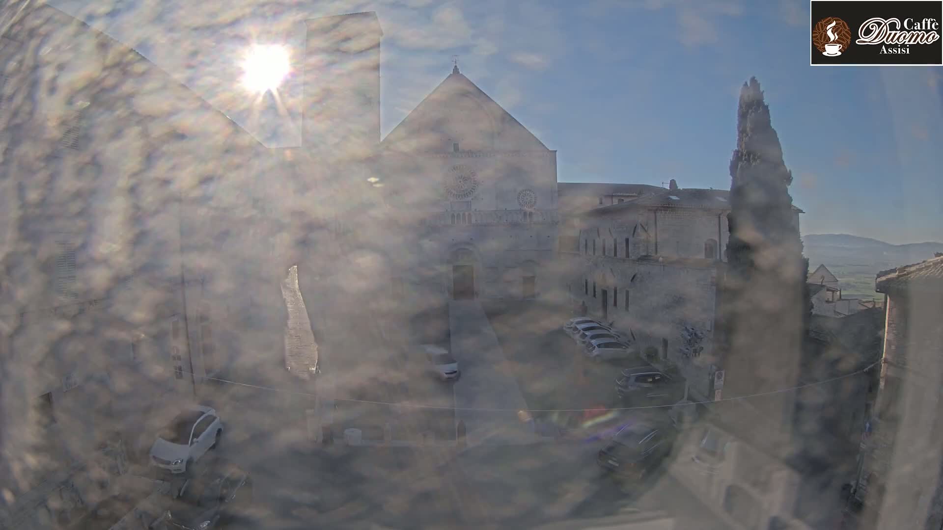 A hazy, sunny view from a window shows a stone courtyard with cars parked, overlooked by a large church and other buildings.