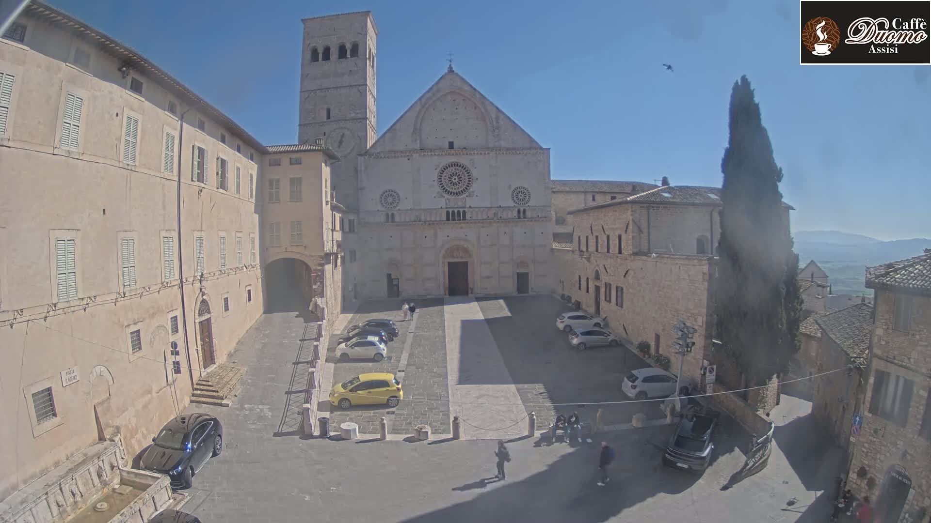 A large stone cathedral sits in a town square with several cars parked and a few pedestrians walking on a sunny day.