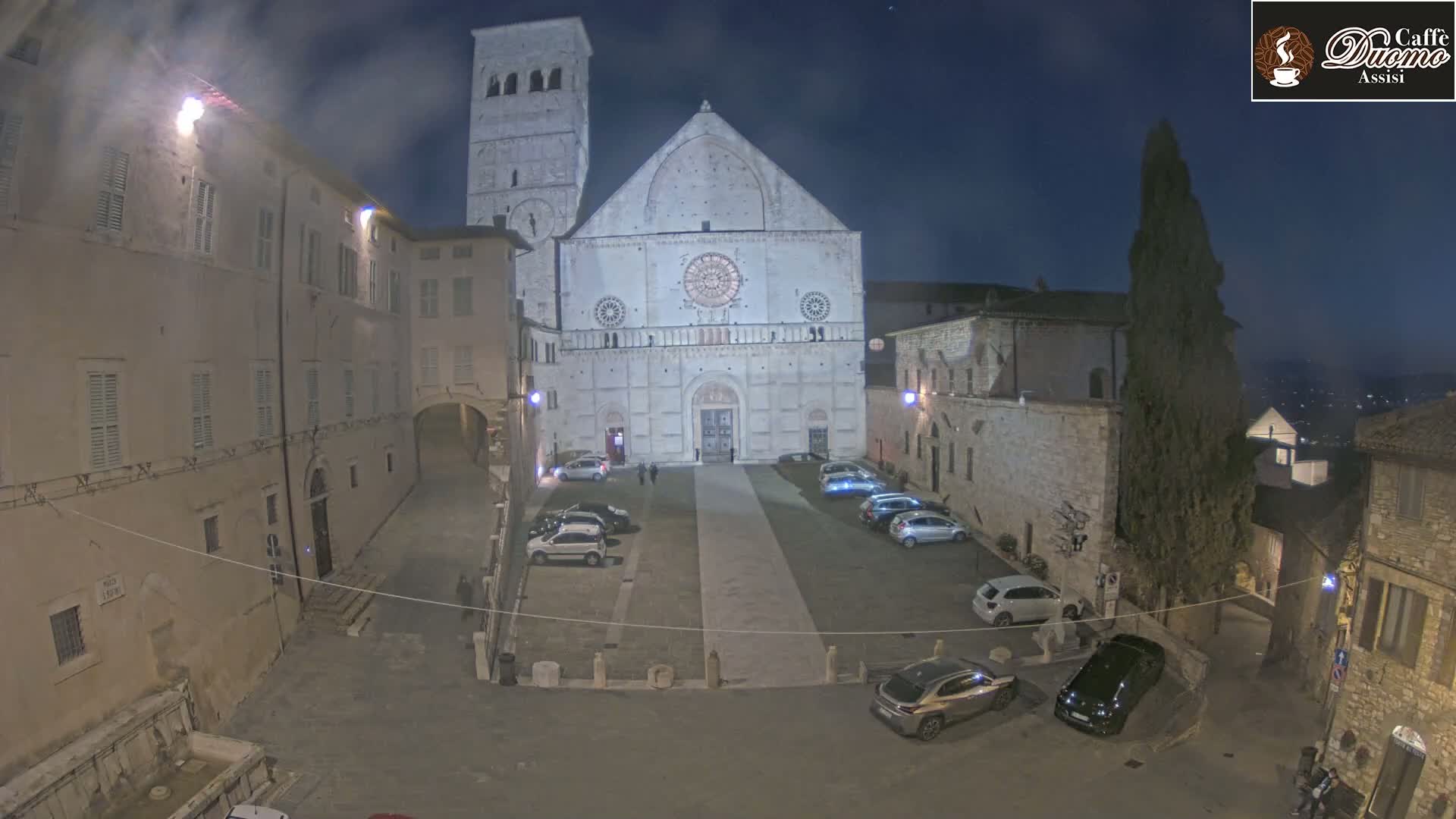 A nighttime, outdoor view of a large stone cathedral in a town square with several cars parked, illuminated by artificial lights.