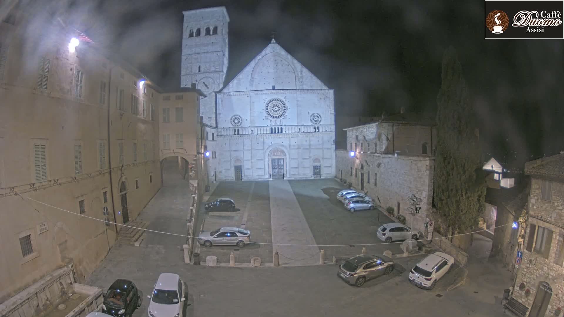 A large stone cathedral is visible in a town square at night under hazy conditions, surrounded by parked cars and older buildings.