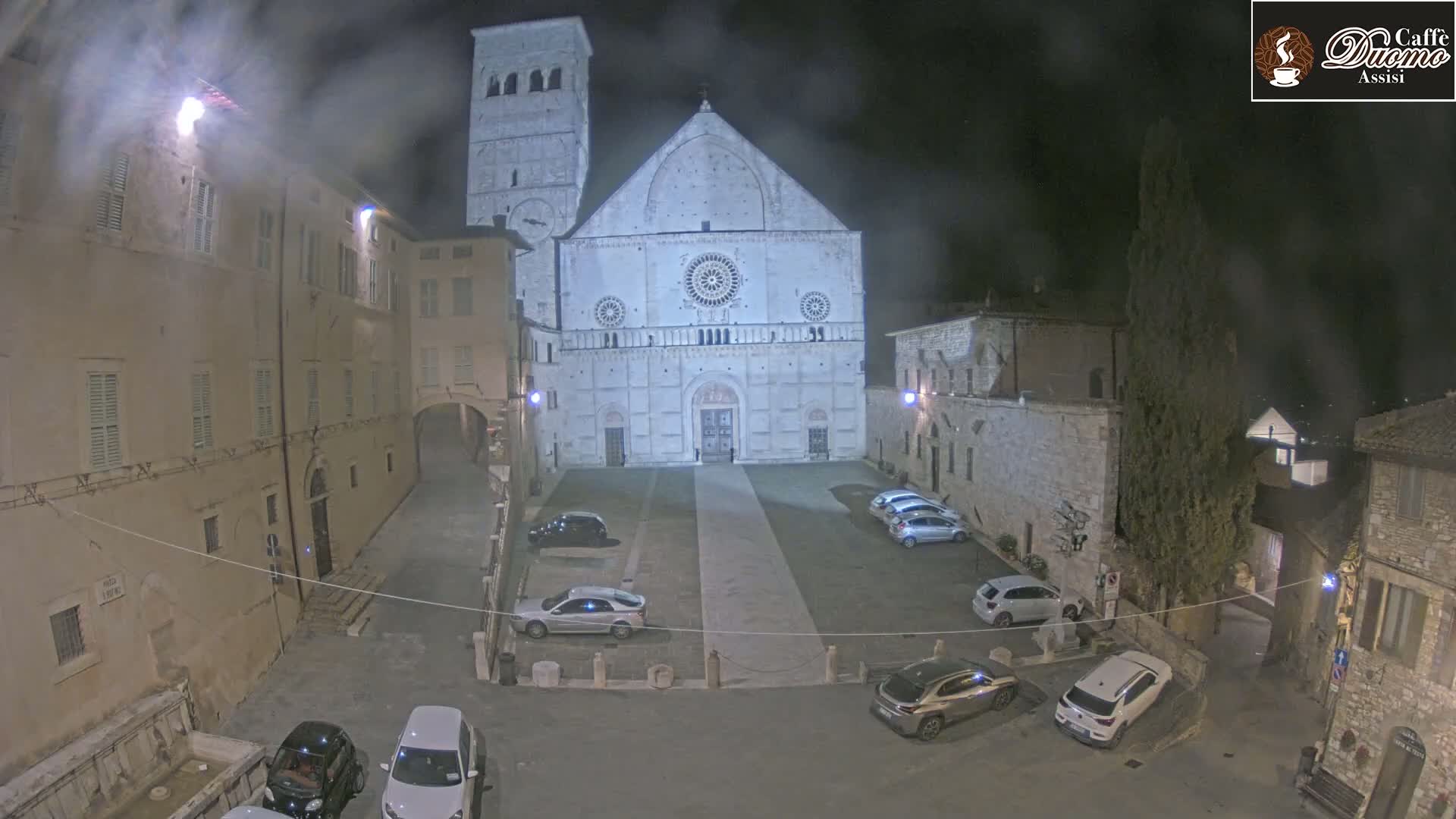 A nighttime, slightly hazy view of a town square with parked cars in front of a large stone cathedral.