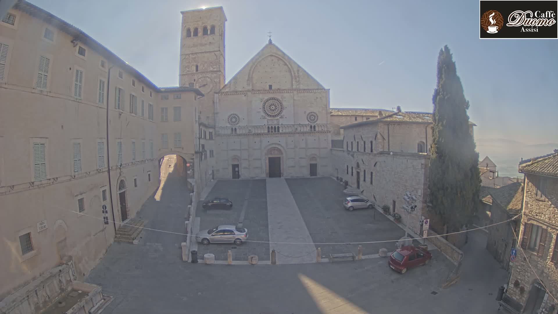 A light-colored stone cathedral and adjacent buildings are visible in a sunny, outdoor square, with several cars parked in the plaza.