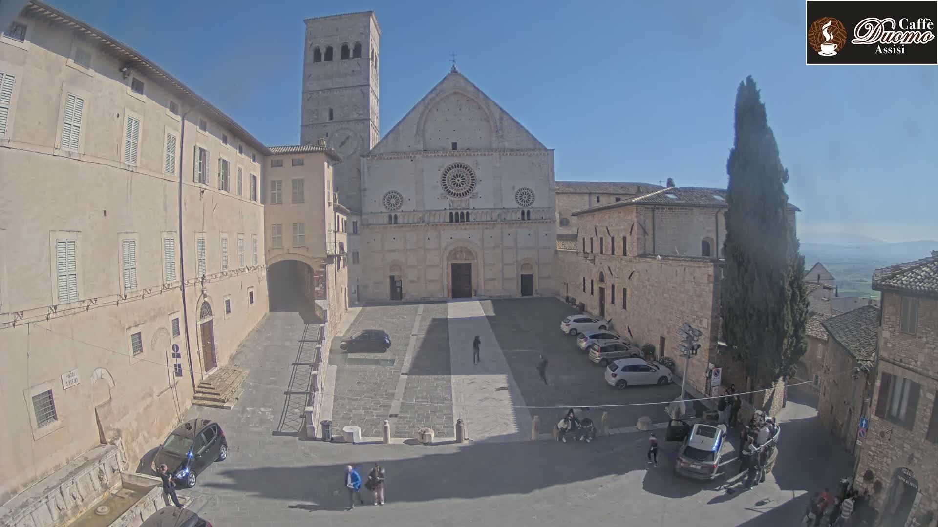 A large stone cathedral sits in a sunny town square, surrounded by other stone buildings and a few parked cars, with several people walking around.