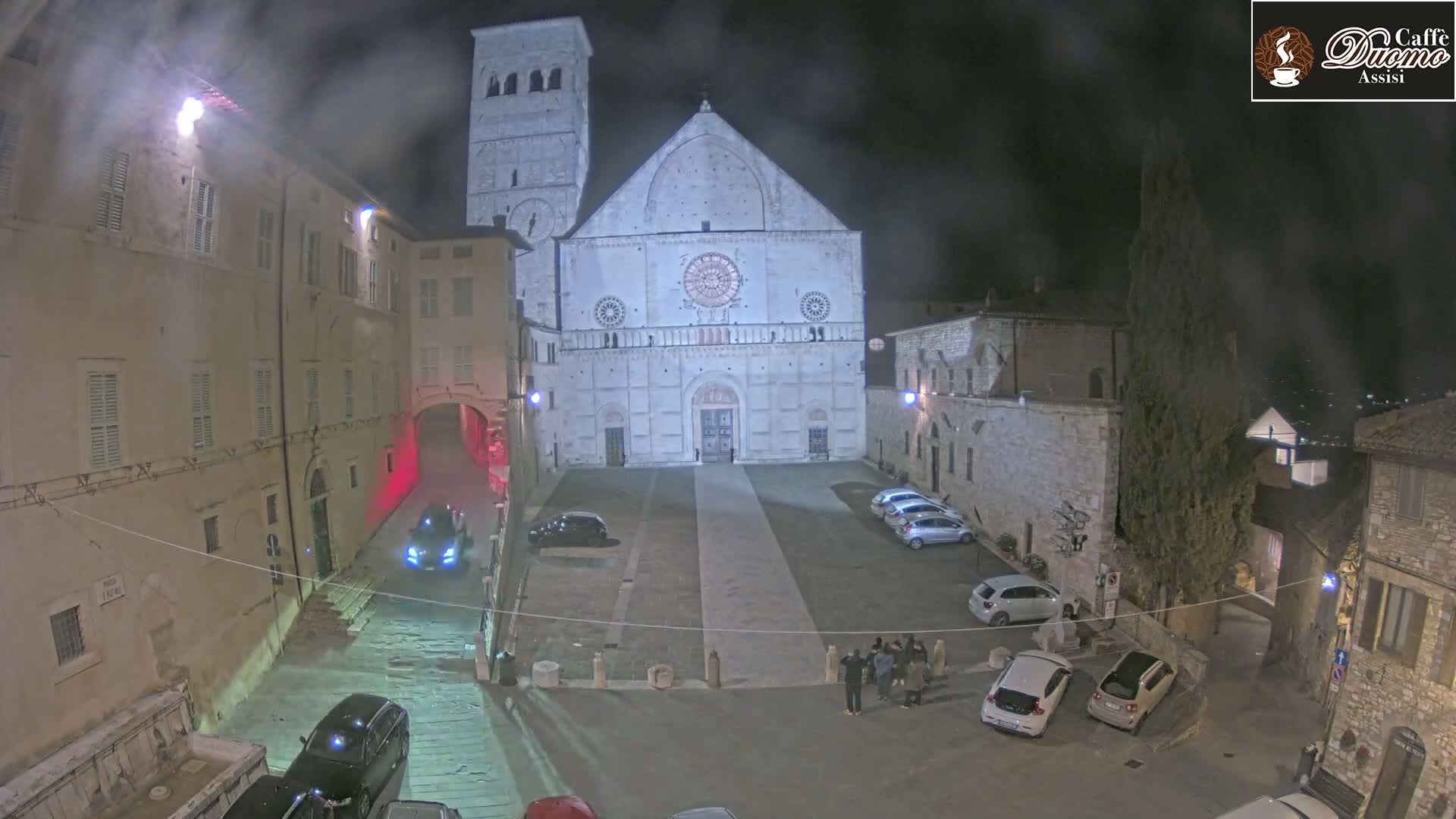 A large, light-colored cathedral stands in a dimly lit, empty town square at night, surrounded by old stone buildings under a hazy sky.