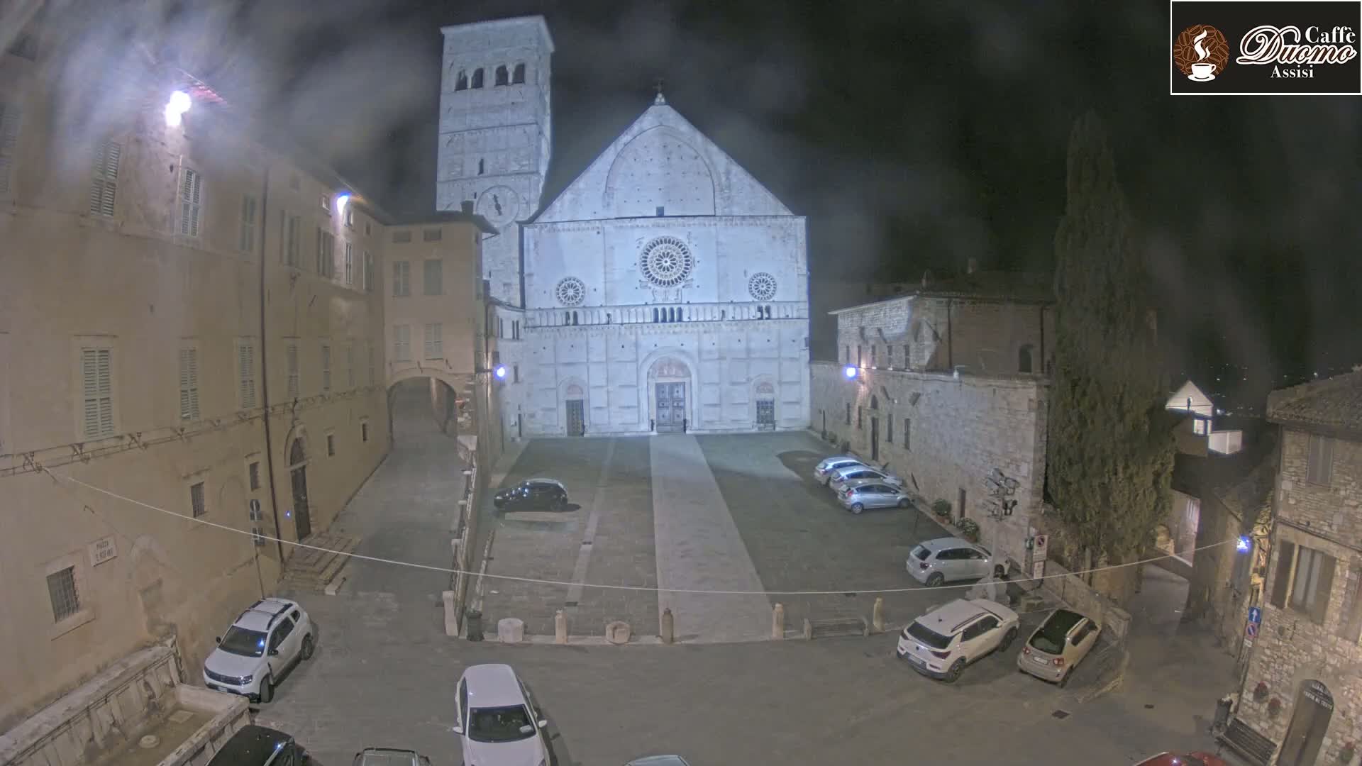 A nighttime view of a cathedral's facade in a town square, with several cars parked, under a cloudy sky.