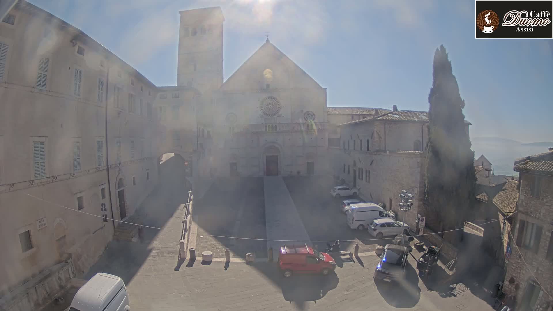 A large stone church is situated in a sunny plaza, surrounded by buildings, with several vehicles parked nearby.