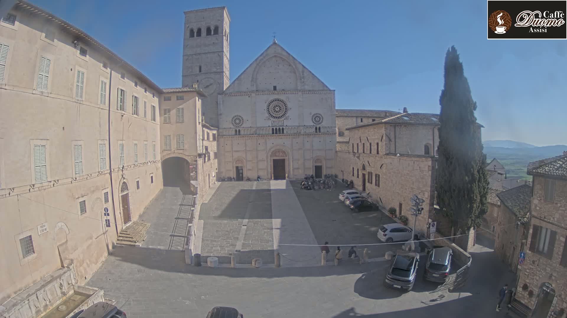 A large stone cathedral sits in a sunny town square, surrounded by other stone buildings and a few parked cars.