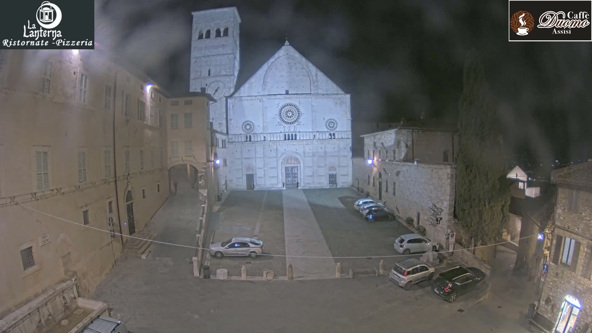 A nighttime, foggy view of a large stone cathedral in a plaza with several cars parked, surrounded by stone buildings.