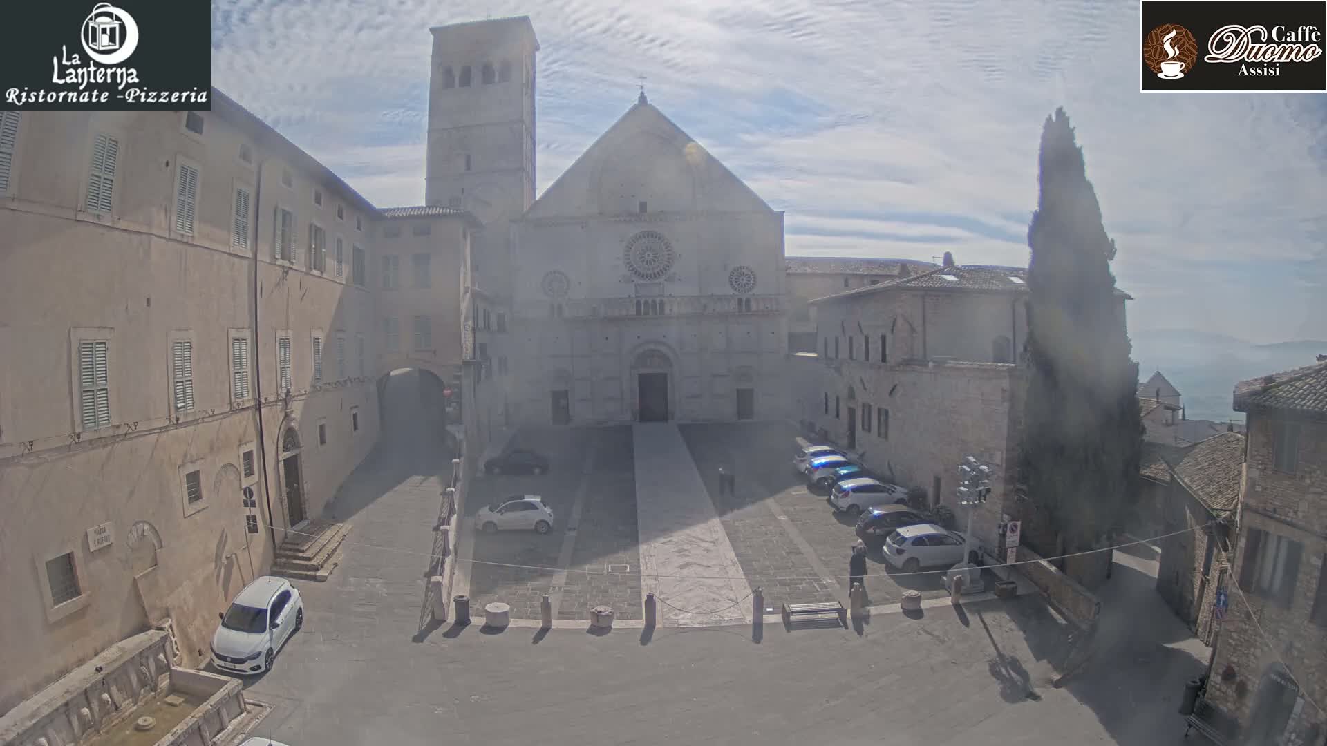 A large stone church stands in a town square on a partly sunny day, with several cars parked nearby and a few pedestrians visible.