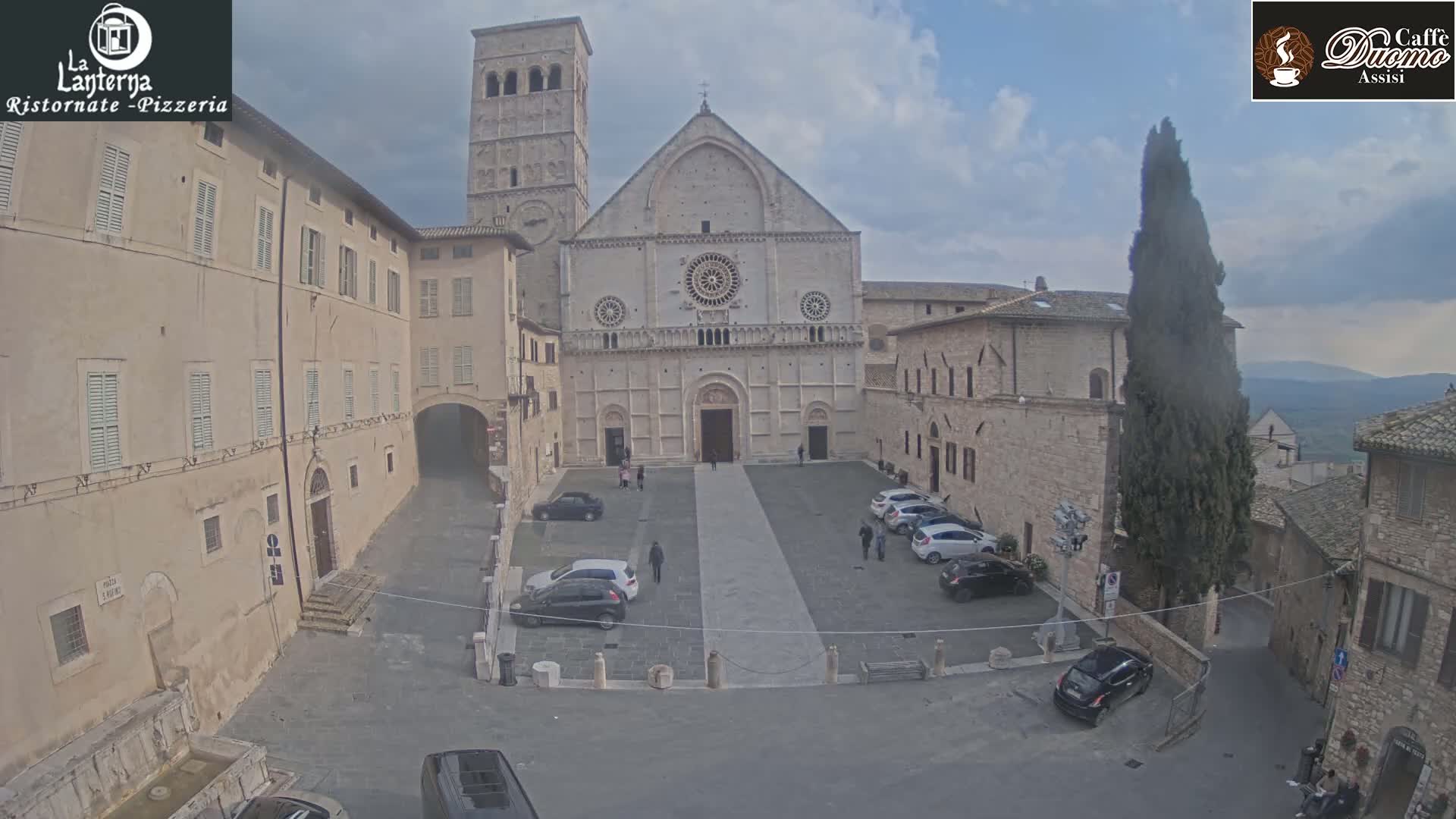 A large stone cathedral sits in a town square with several parked cars and a few pedestrians, under an overcast sky.