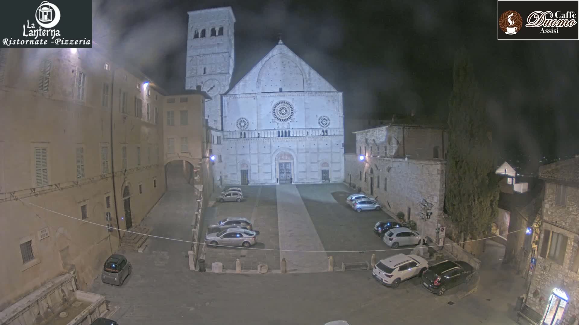 A nighttime view of a cathedral's facade and surrounding plaza with several cars parked in the square under hazy conditions.
