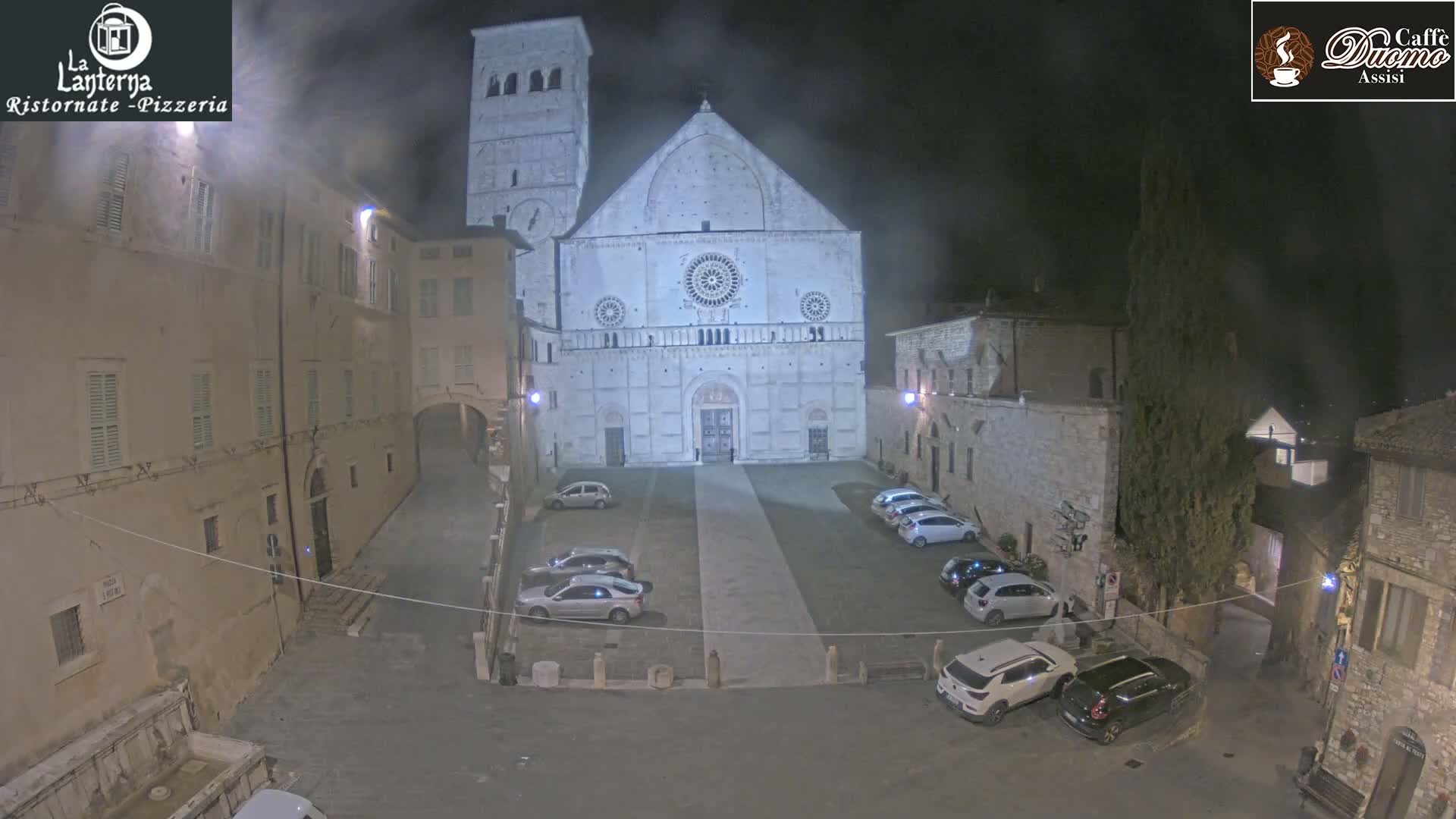 A nighttime view of a town square with a large stone cathedral at its center, surrounded by buildings and parked cars, under a hazy or foggy sky.