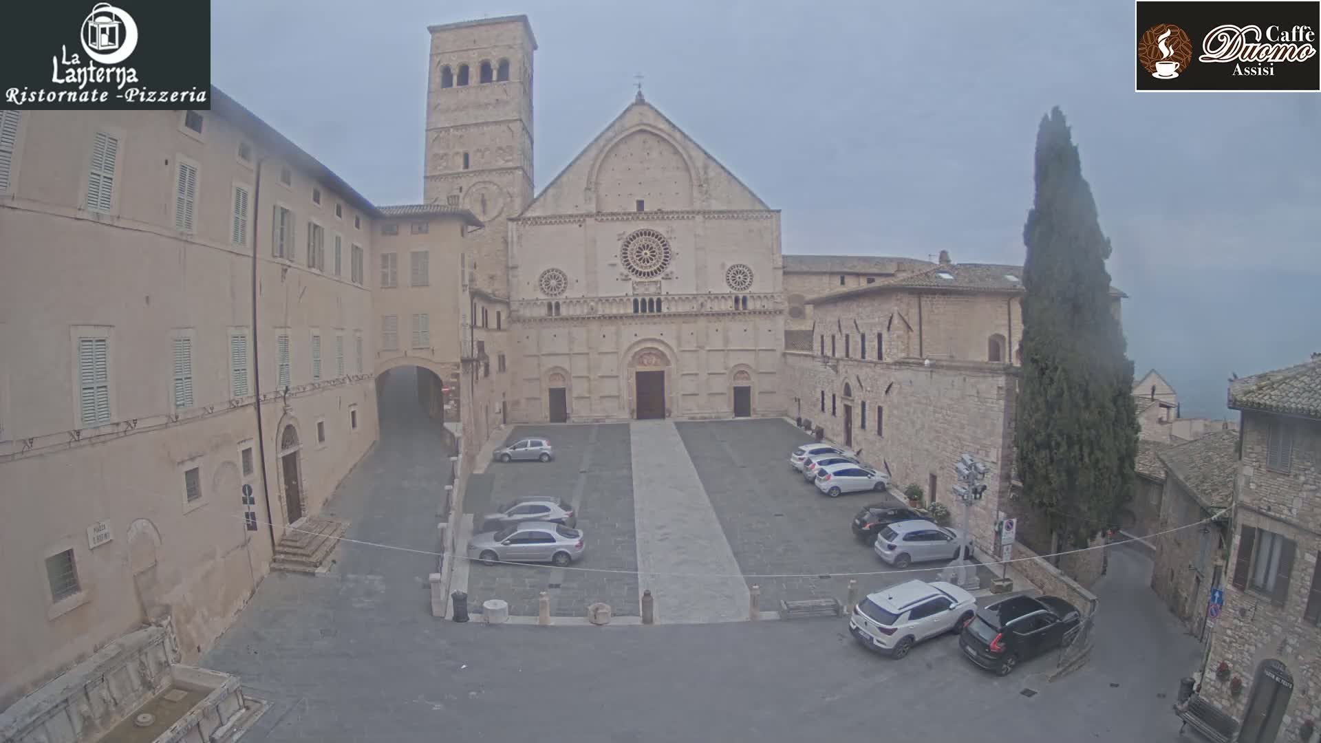 A stone plaza with several parked cars sits in front of a large cathedral on an overcast day.