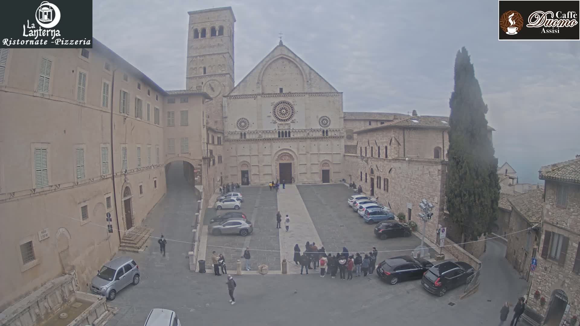 A large stone cathedral in a town square, surrounded by buildings, on an overcast day with a few cars parked.