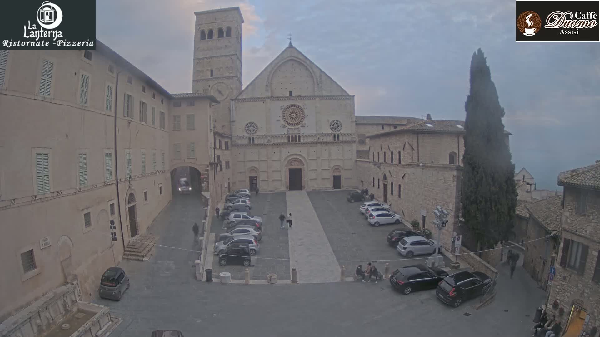 A large stone church and surrounding buildings, a parking lot with several cars, and a few pedestrians are visible in a town square on an overcast day.
