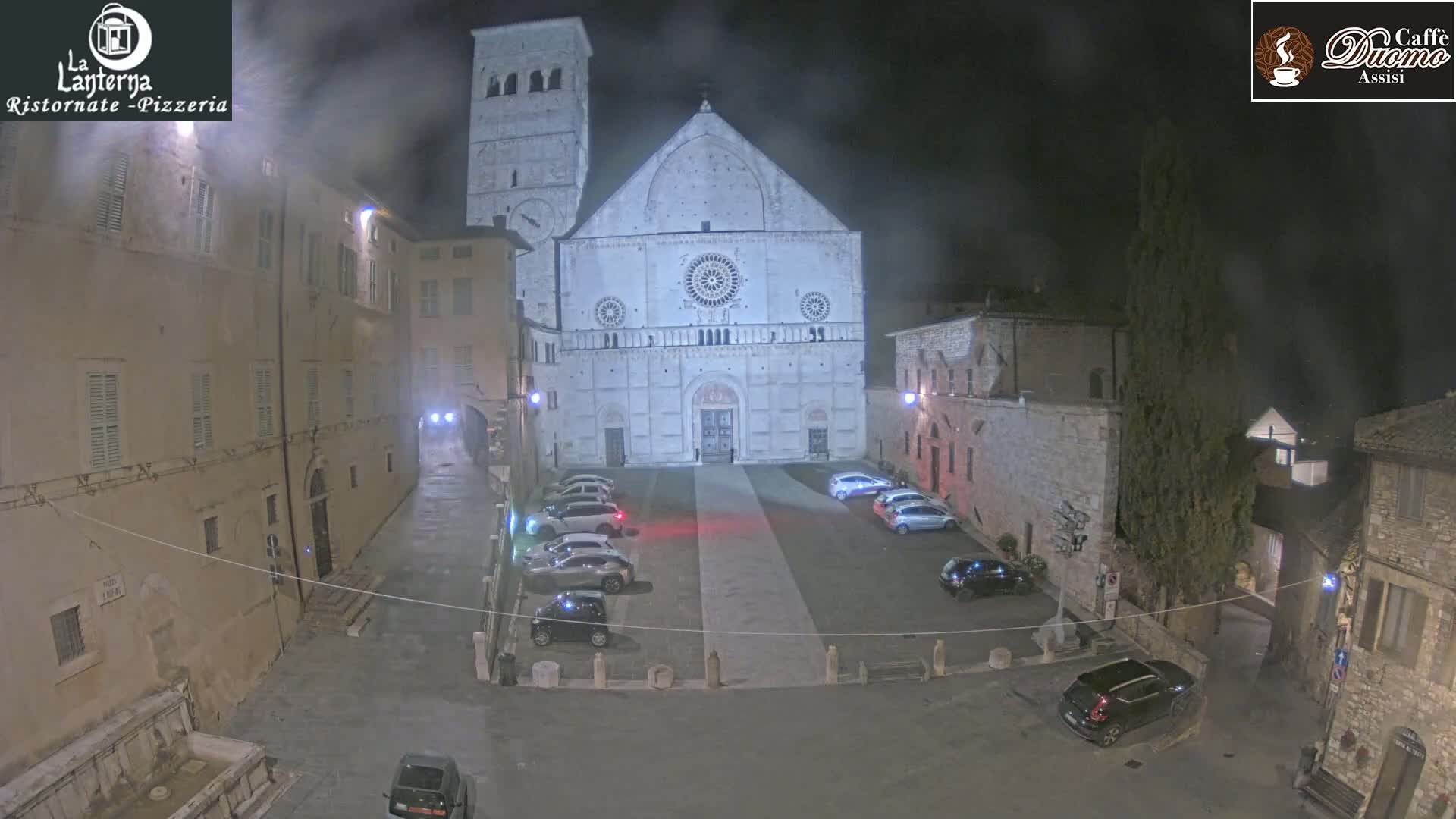 A large stone cathedral stands in a town square at night, under hazy conditions, with several cars parked in the square.