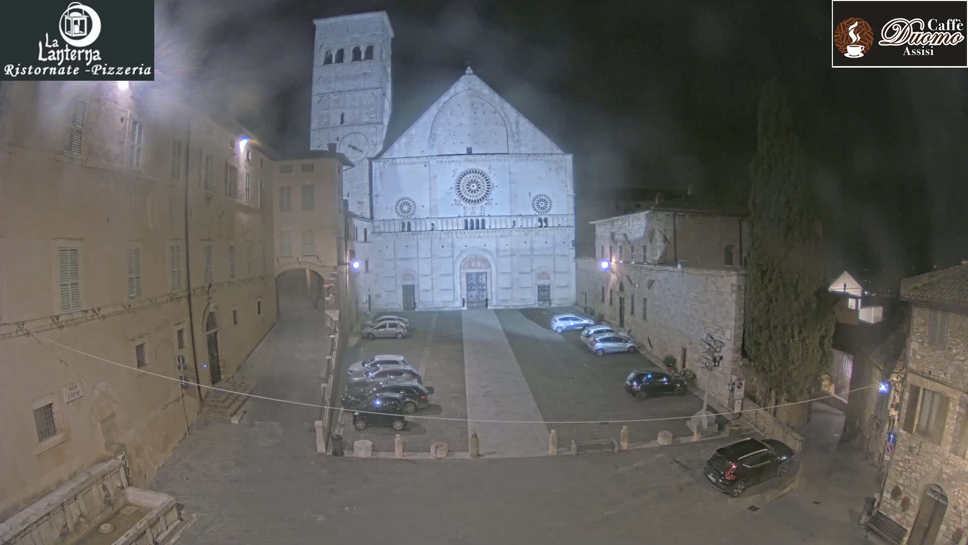 A nighttime view of a town square with a large stone cathedral at the center, several cars parked in the square, and older buildings surrounding the area, under hazy or foggy conditions.