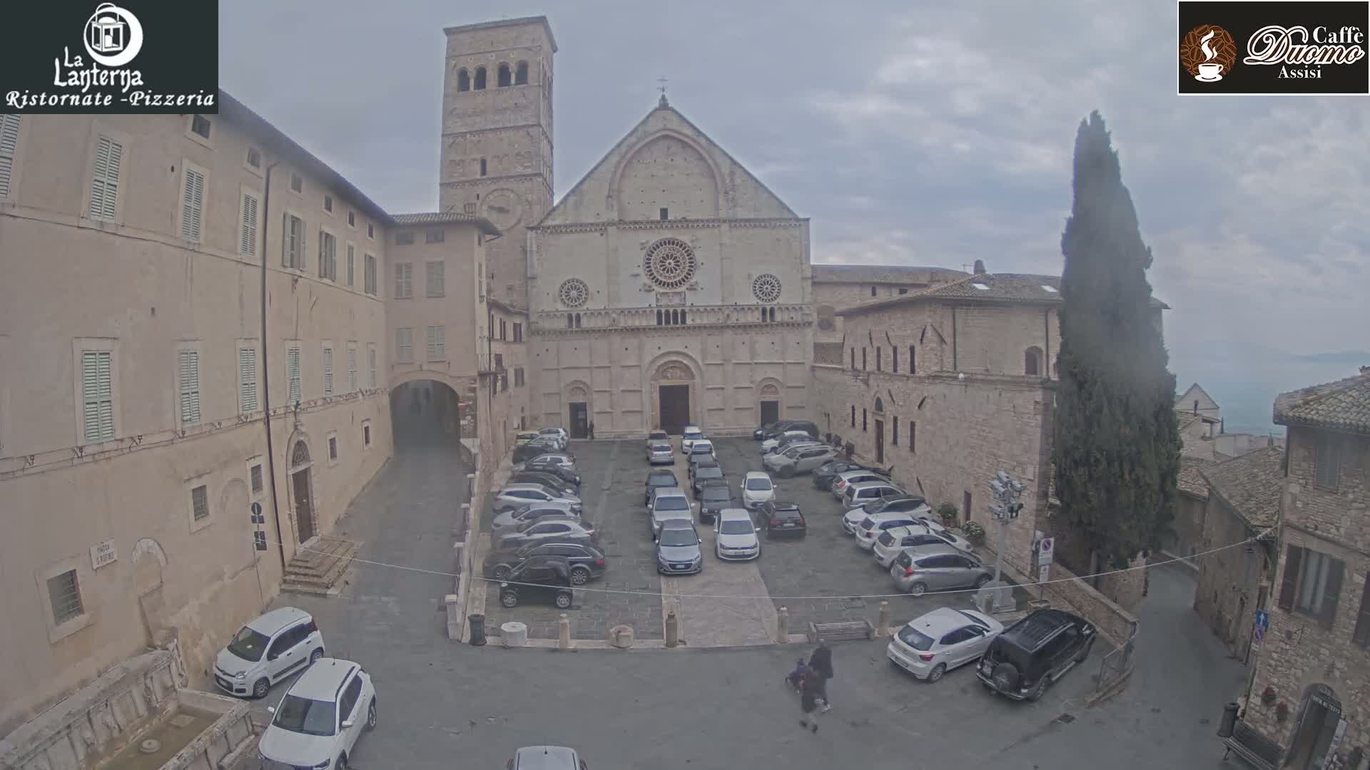 A stone cathedral and surrounding buildings are visible in an overcast parking lot with several cars parked.