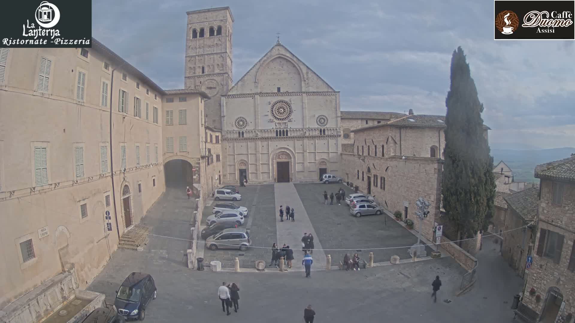 A large stone church stands in a plaza surrounded by buildings under an overcast sky, with several people and parked cars visible.
