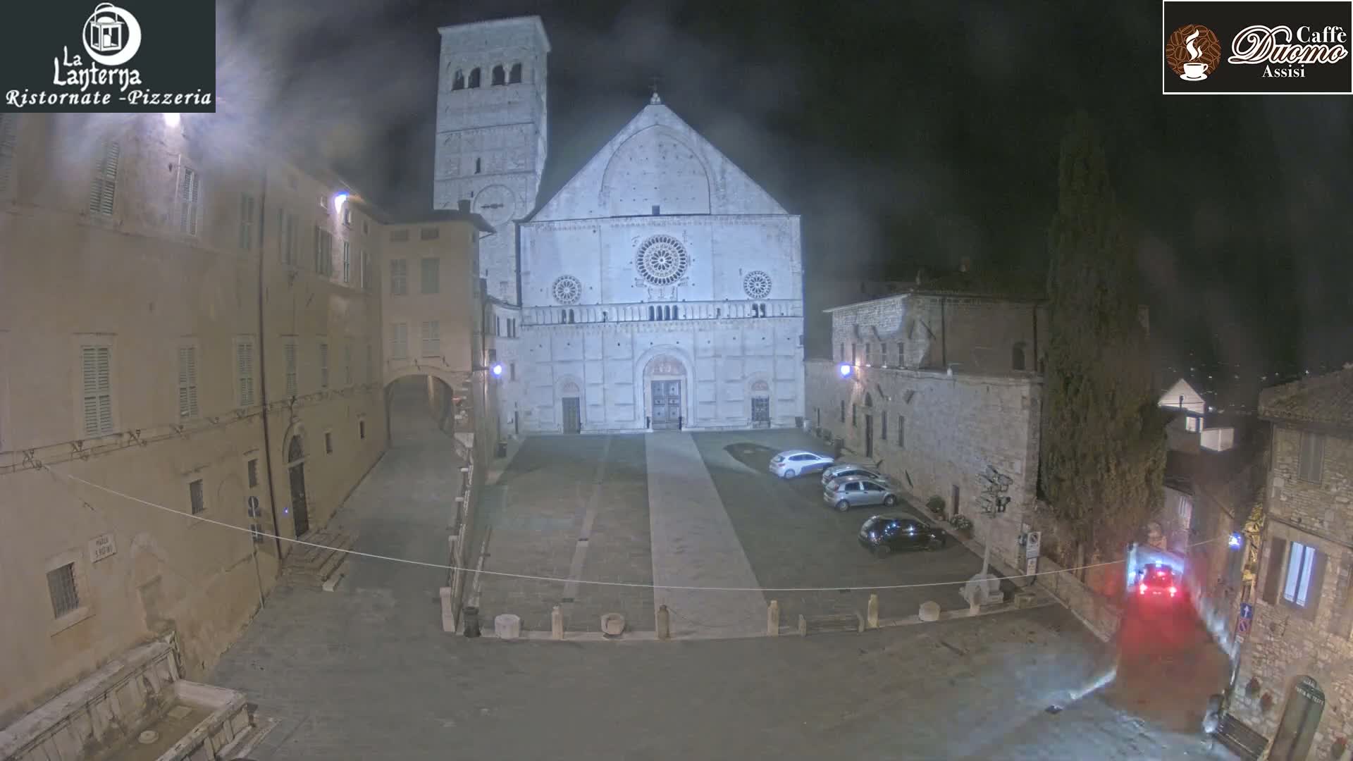 A nighttime view of a town square, featuring a large cathedral illuminated against a hazy sky, with several cars parked nearby.