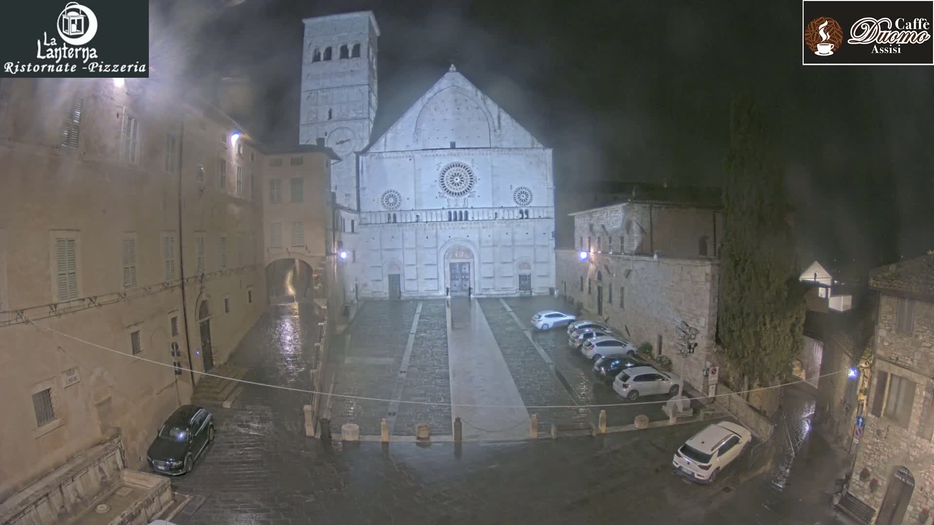 A large cathedral stands in a town square at night during a light rain, surrounded by old stone buildings and parked cars.