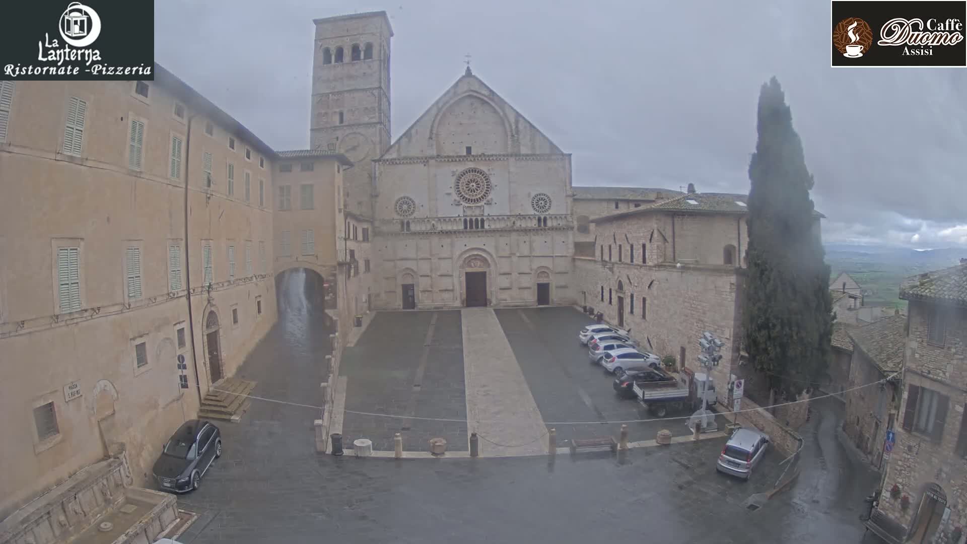 A large stone church sits in a town square on a rainy day, surrounded by other stone buildings and a few parked cars.