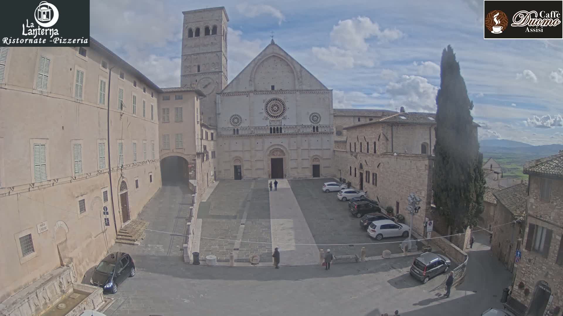 A large stone cathedral sits in a town square on a partly cloudy day, surrounded by other stone buildings and cars parked in the square.