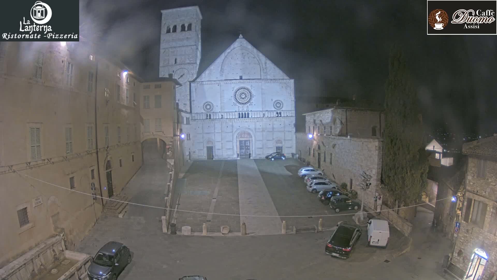 A nighttime view of a town square with a large stone cathedral at the center, several cars parked nearby, and buildings lining the square under hazy, foggy conditions.