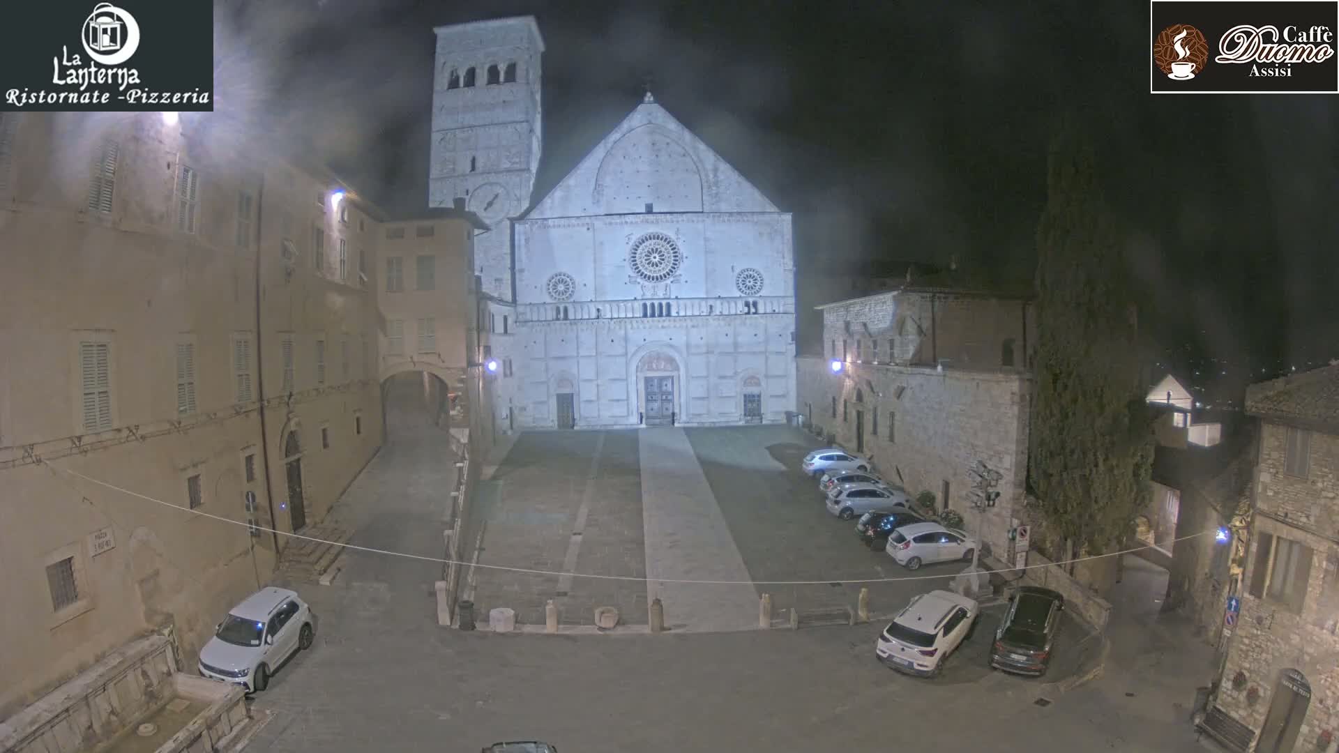 A nighttime view of a large stone cathedral in a town square, with several cars parked nearby under hazy or foggy conditions.