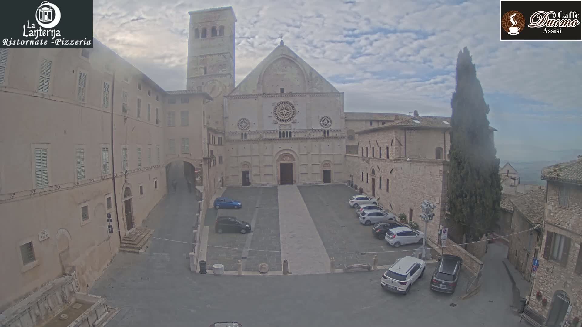 A stone cathedral is visible in a town square with overcast skies, several cars parked nearby, and surrounding stone buildings.