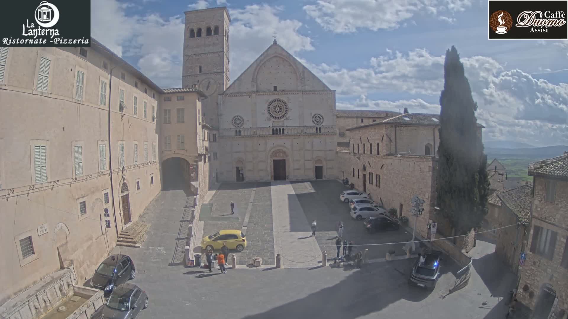 A partly cloudy day reveals a large stone cathedral in a town square, surrounded by old stone buildings and a few parked cars.