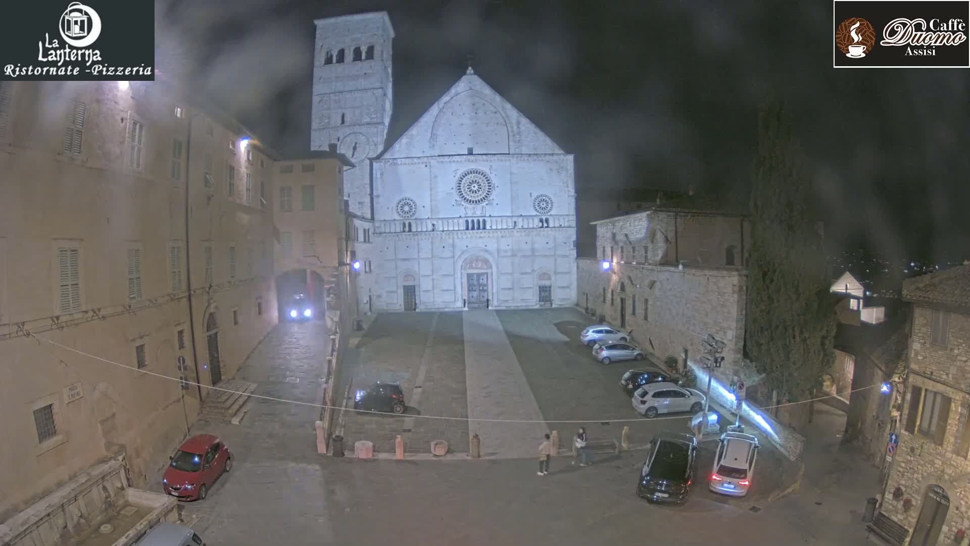 A nighttime view of a town square with a large cathedral in the background, under hazy or foggy conditions.