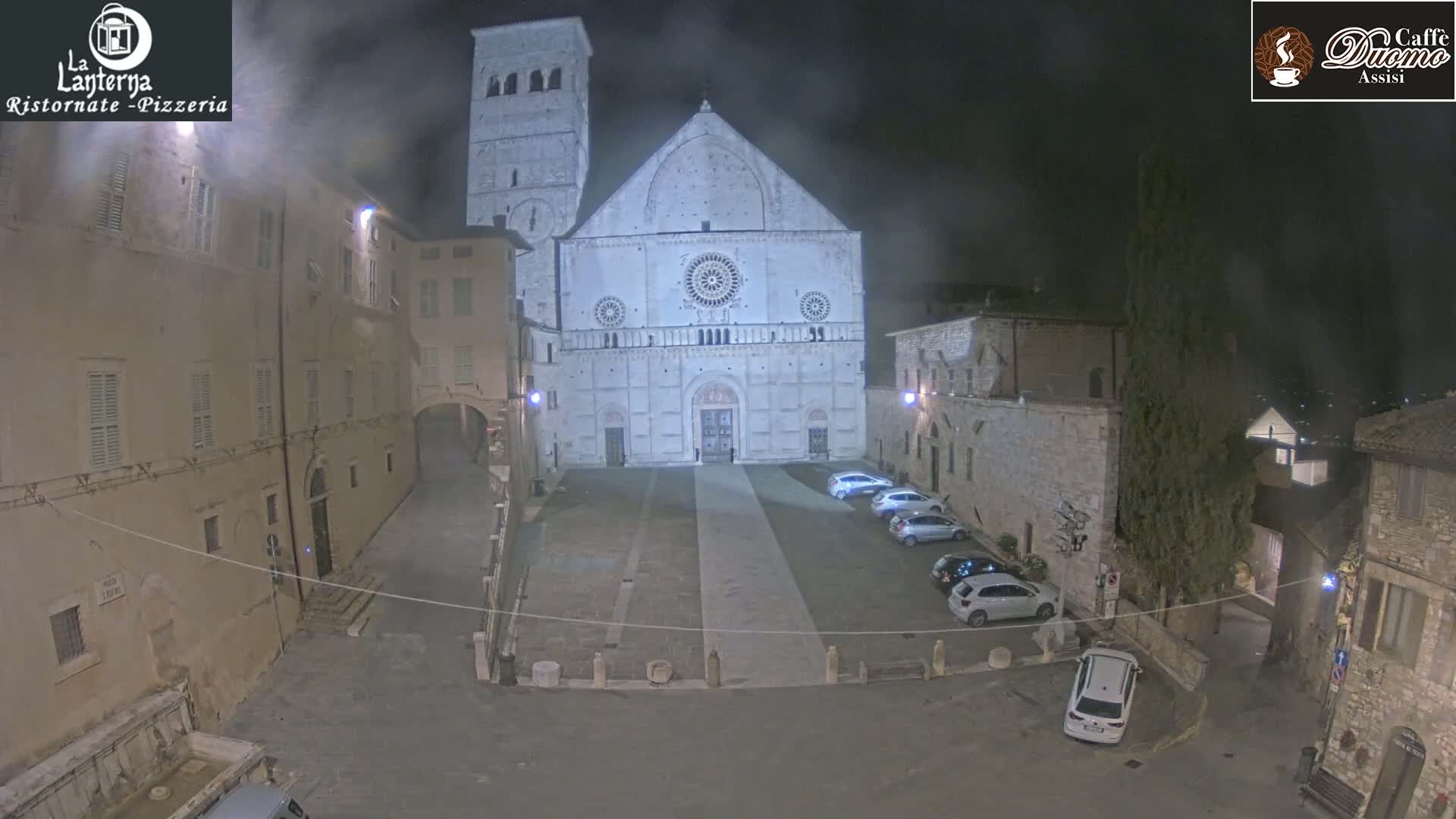 A nighttime view of a stone plaza with parked cars in front of a large cathedral under a hazy sky.