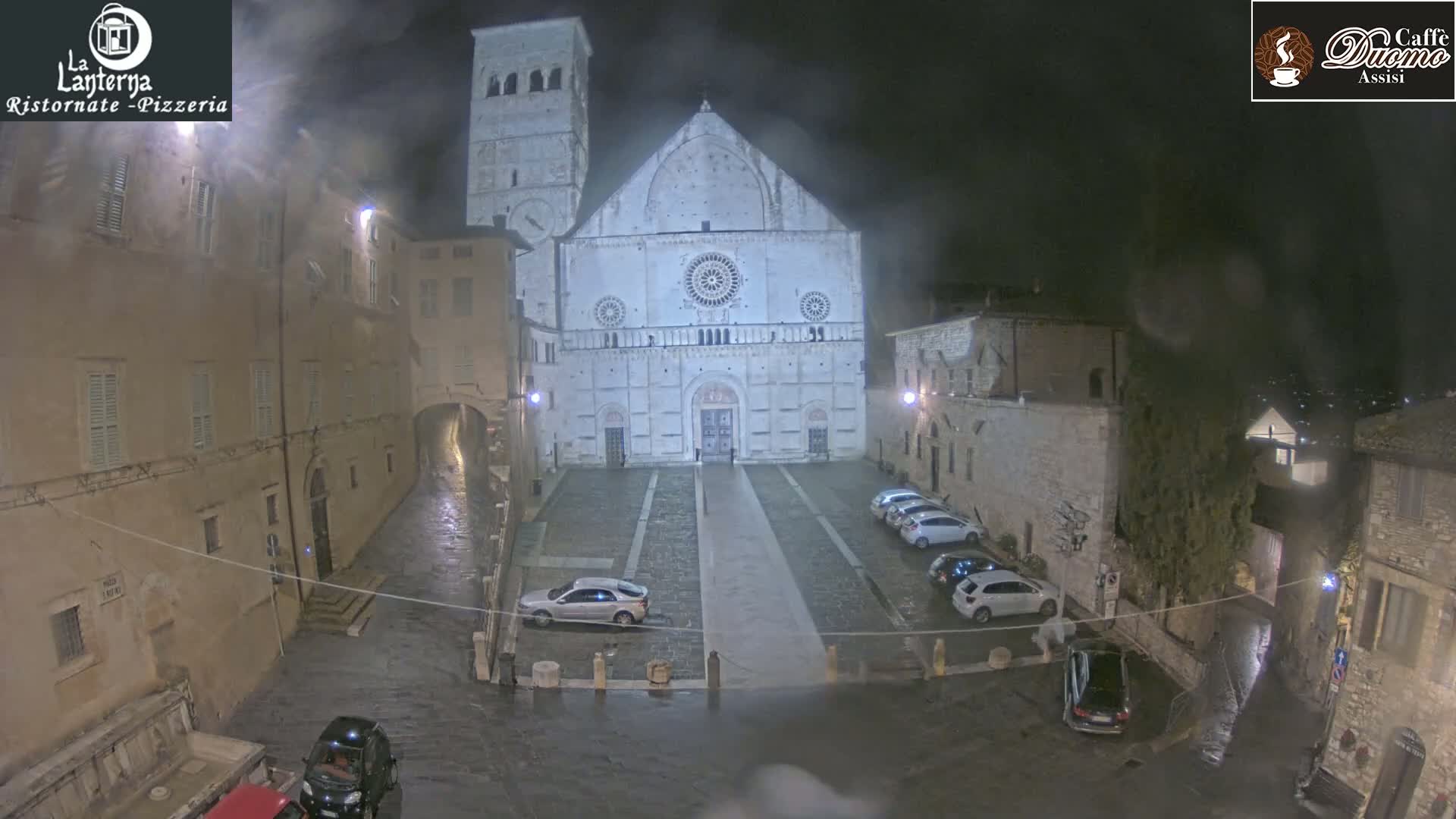 A large stone cathedral is the focal point of a rain-soaked town square at night, with several cars parked nearby and the surrounding buildings dimly lit.