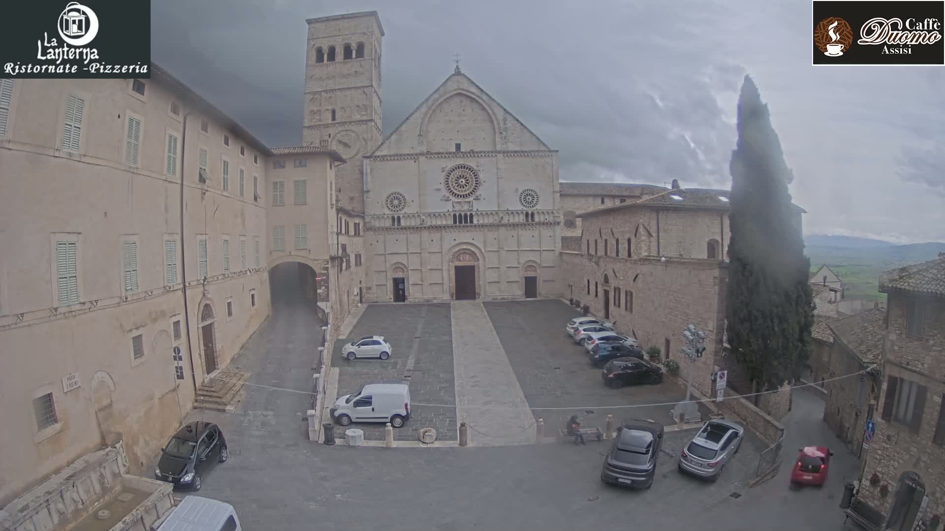A large, light-colored stone church stands in a square with several cars parked, under an overcast sky.